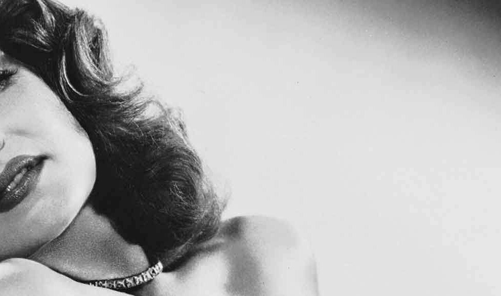 A black and white picture of Rita Hayworth in a black, off the shoulder dress, looking very sultry and glamorous.